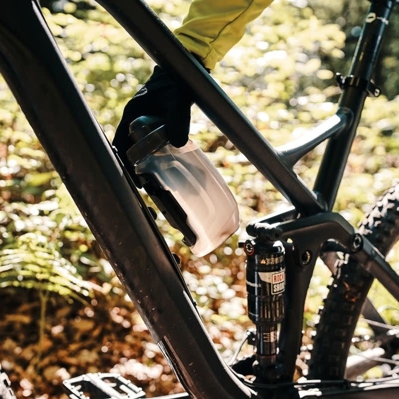 Someone reaching down to attach a bottle to a mount on a dual suspension mountain bike, with forest in the background