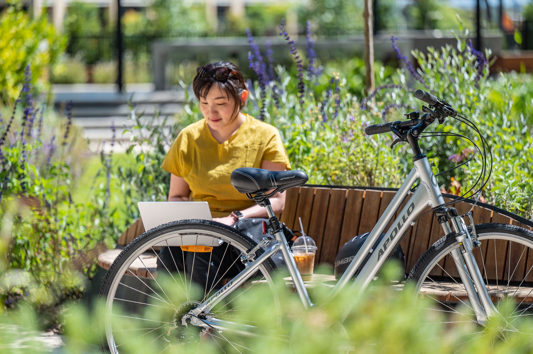 Person sitting on a bench with an Apollo step through hybrid bicycle, using a laptop in a garden setting