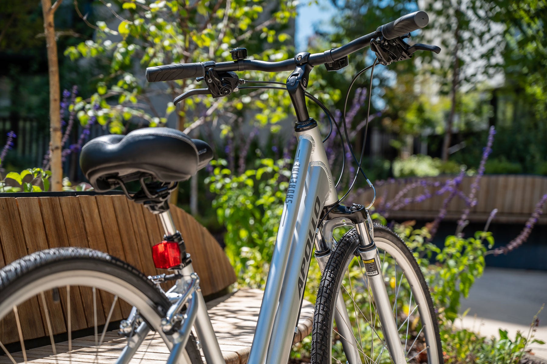 A silver Apollo Step Through Hybrid bicycle with black seat and handlebars parked outdoors with greenery in the background