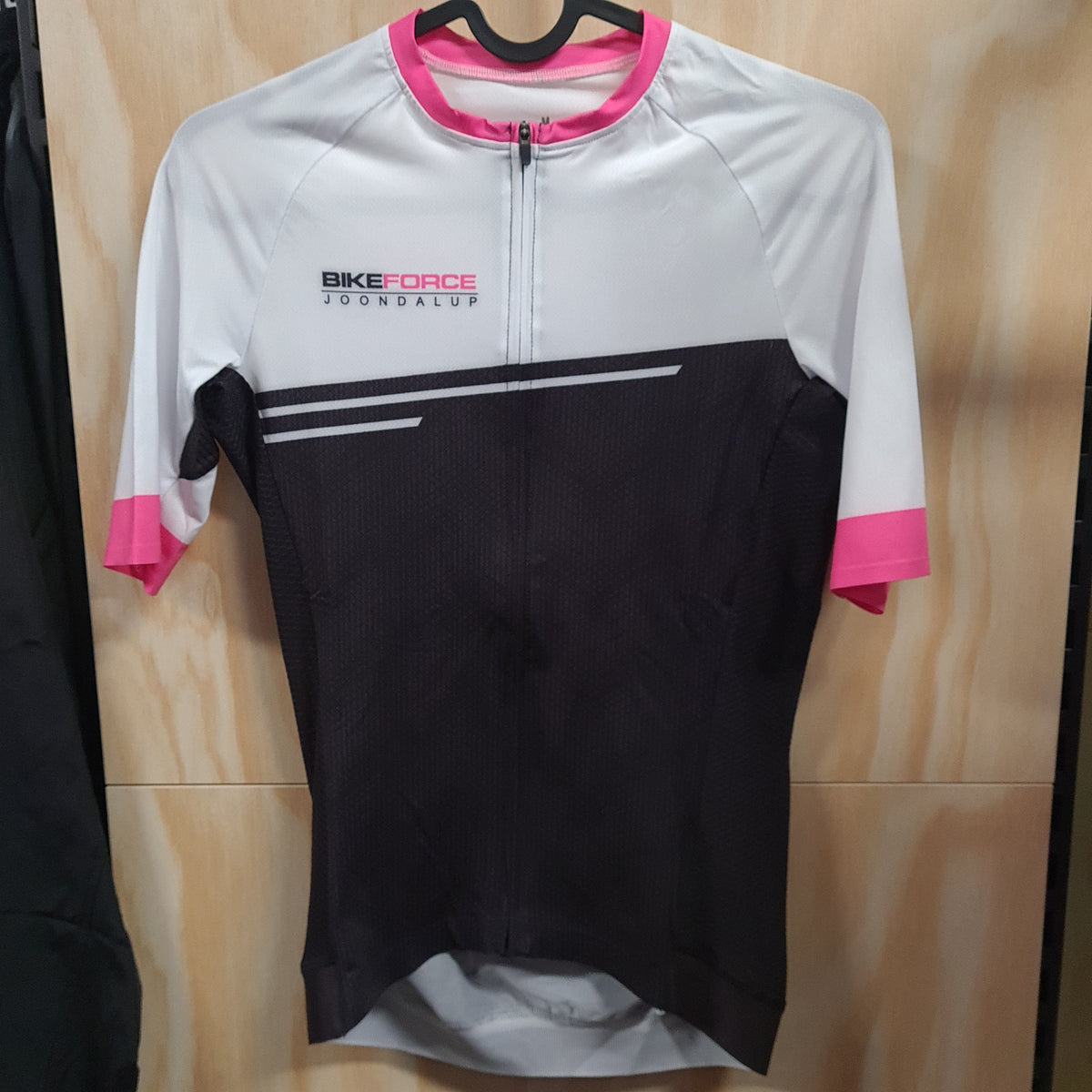 A short-sleeve cycling jersey hanging on a black hanger against a light wood panel background. The jersey has a white upper section, a black lower section, and bright pink accents on the collar, sleeve cuffs, and side panels. The logo 'Bike Force Joondalup' is printed in black and pin on the right upper chest.