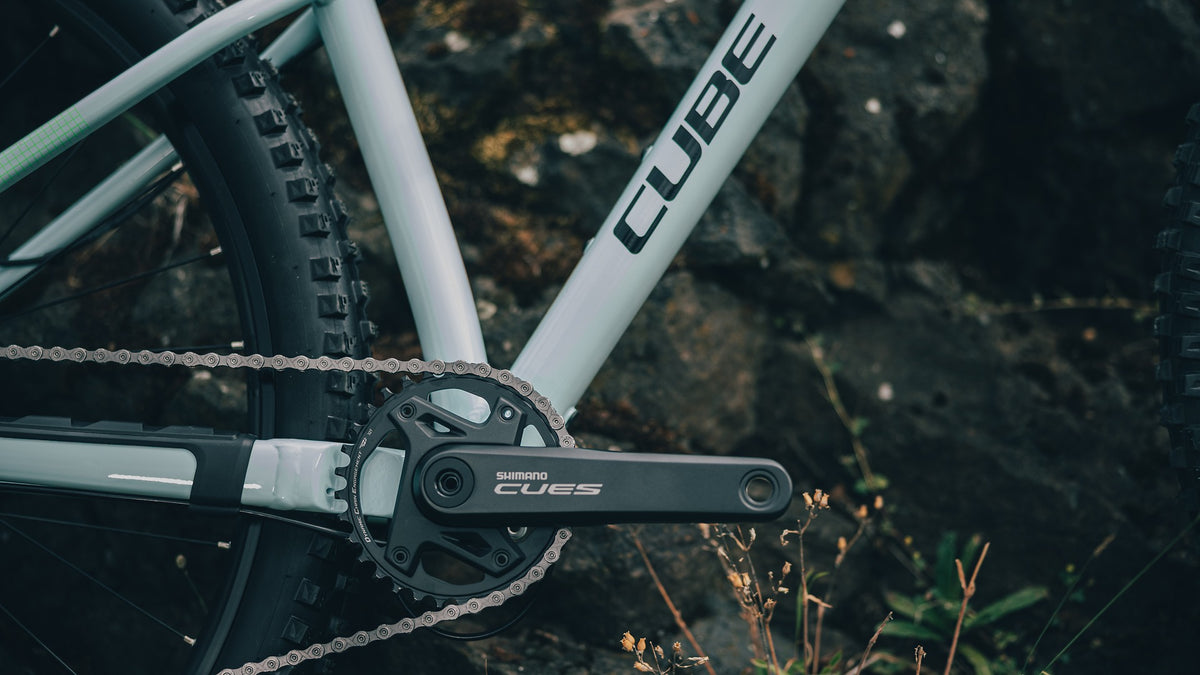 Close-up of a bicycle crankset with a Shimano Cues chainring and a light blue frame labeled CUBE, photographed outdoors against dark rocks and grass.
