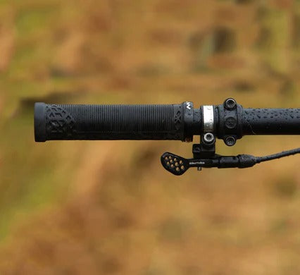 Close-up of a black bicycle handlebar with a textured grip, clamp hardware, and a brake lever, set against a blurred outdoor background.