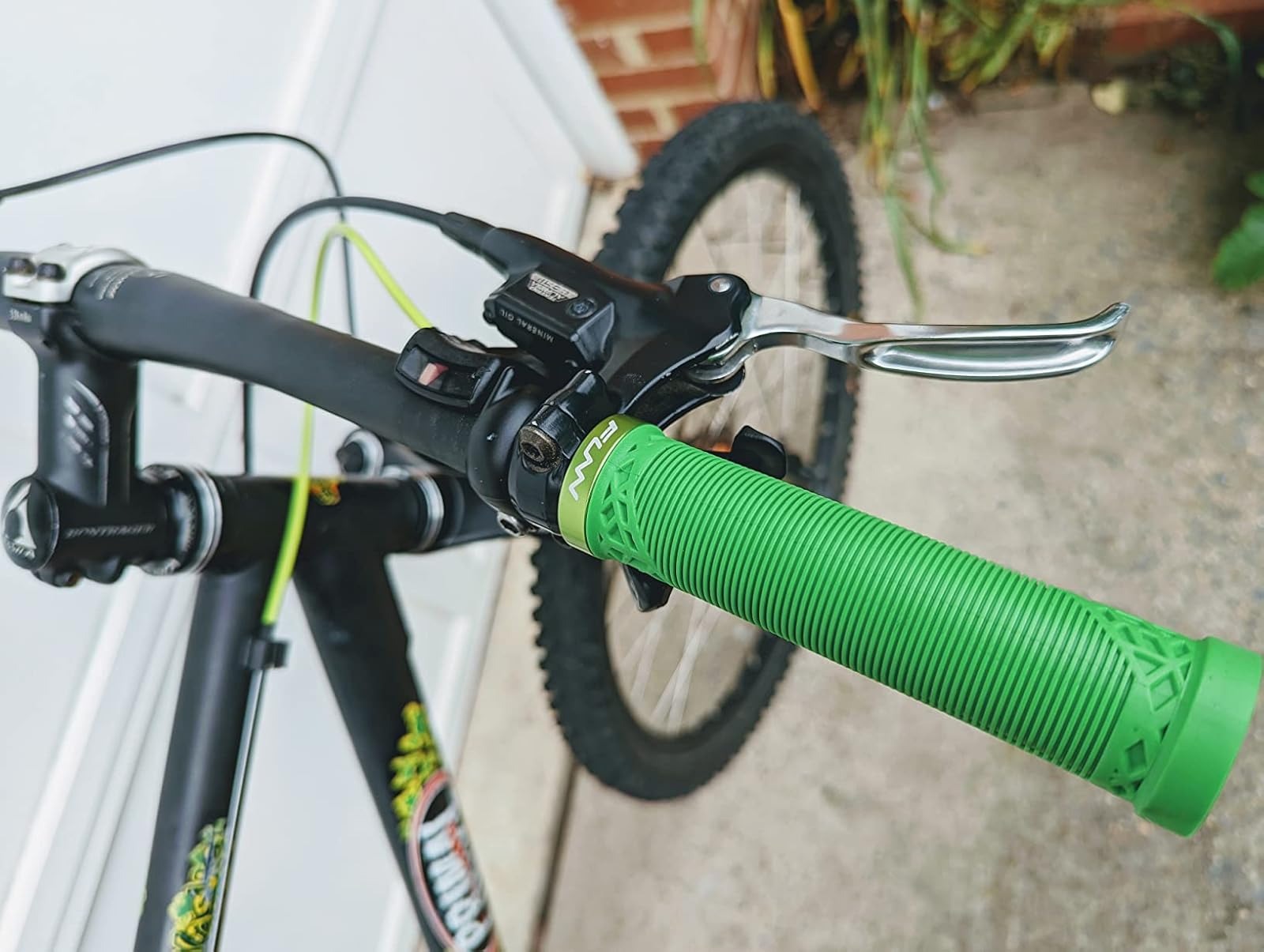 Close-up of a bicycle handlebar with a bright green textured grip and silver brake lever, with part of the black frame and rear wheel visible in an outdoor setting.