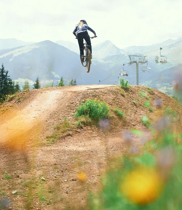 Person on a bicycle performing a jump in a mountainous area with ski lifts in the background.