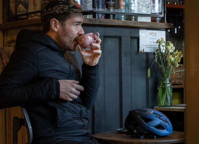 A picture of a man drinking coffee with a Lazer Tonic Kineticore Blue Helmet on the table next to him