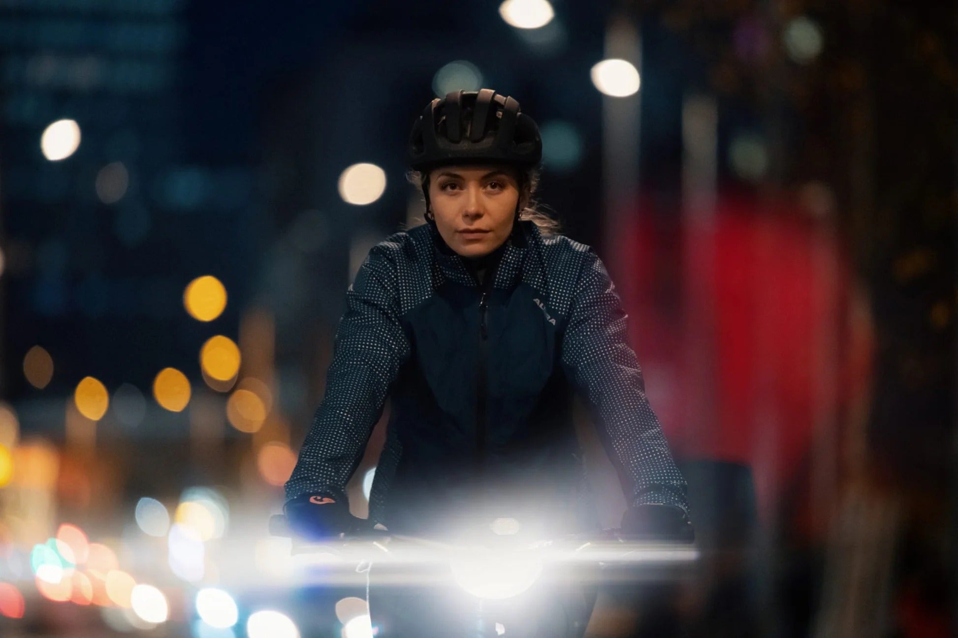 Person riding a bicycle at night with a front light shining against a blurred city lights in the background