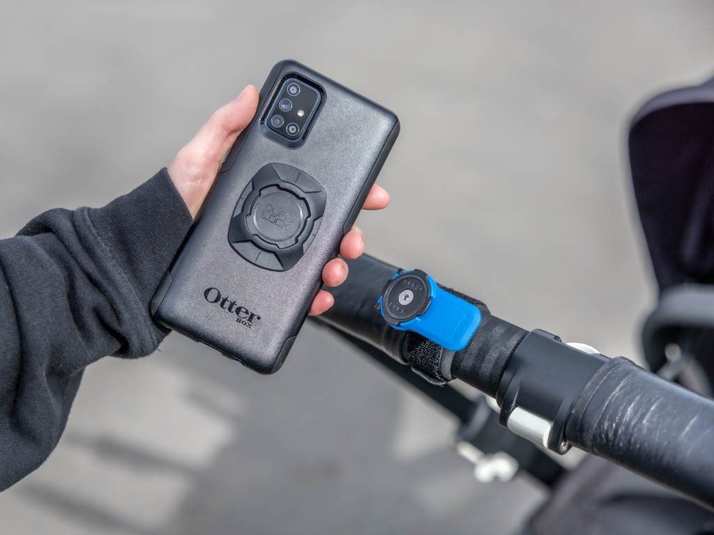 Person holding a black phone case with Quad Lock branding on a blurred background