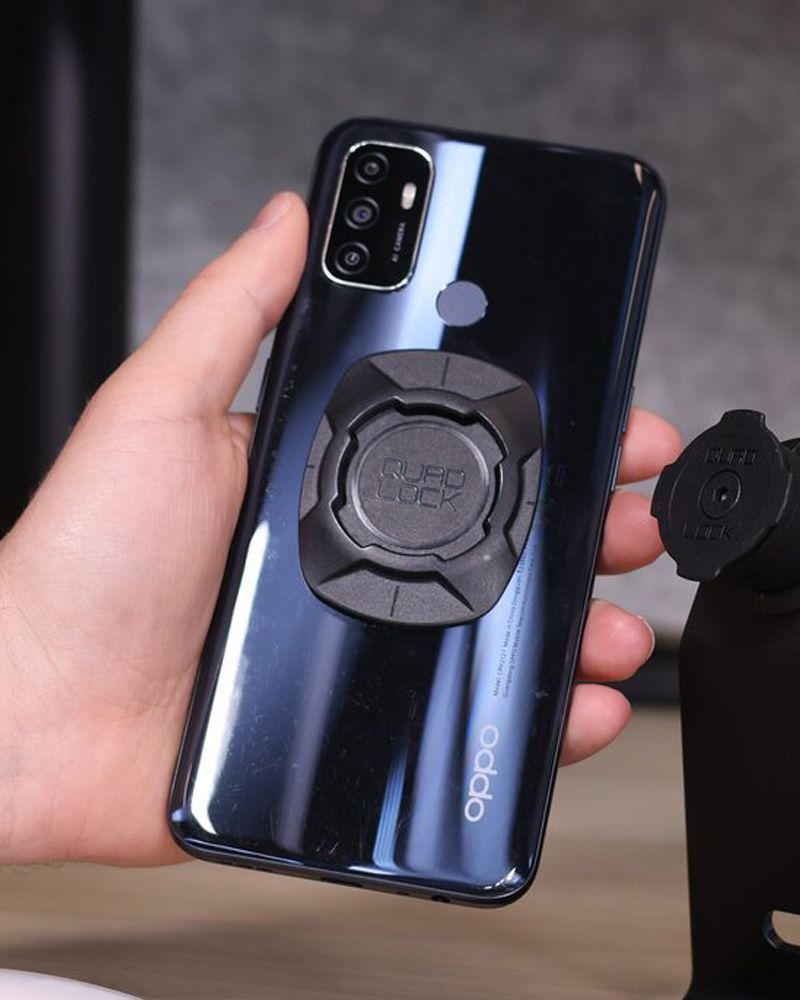 A hand holding a phone with a Quad Lock Phone Mount Adaptor on the back on a blurred background