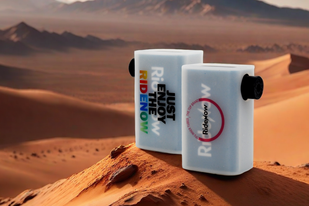 Two white rectangular RideNow electric pumps standing on a desert sand dune with mountains in the background.
