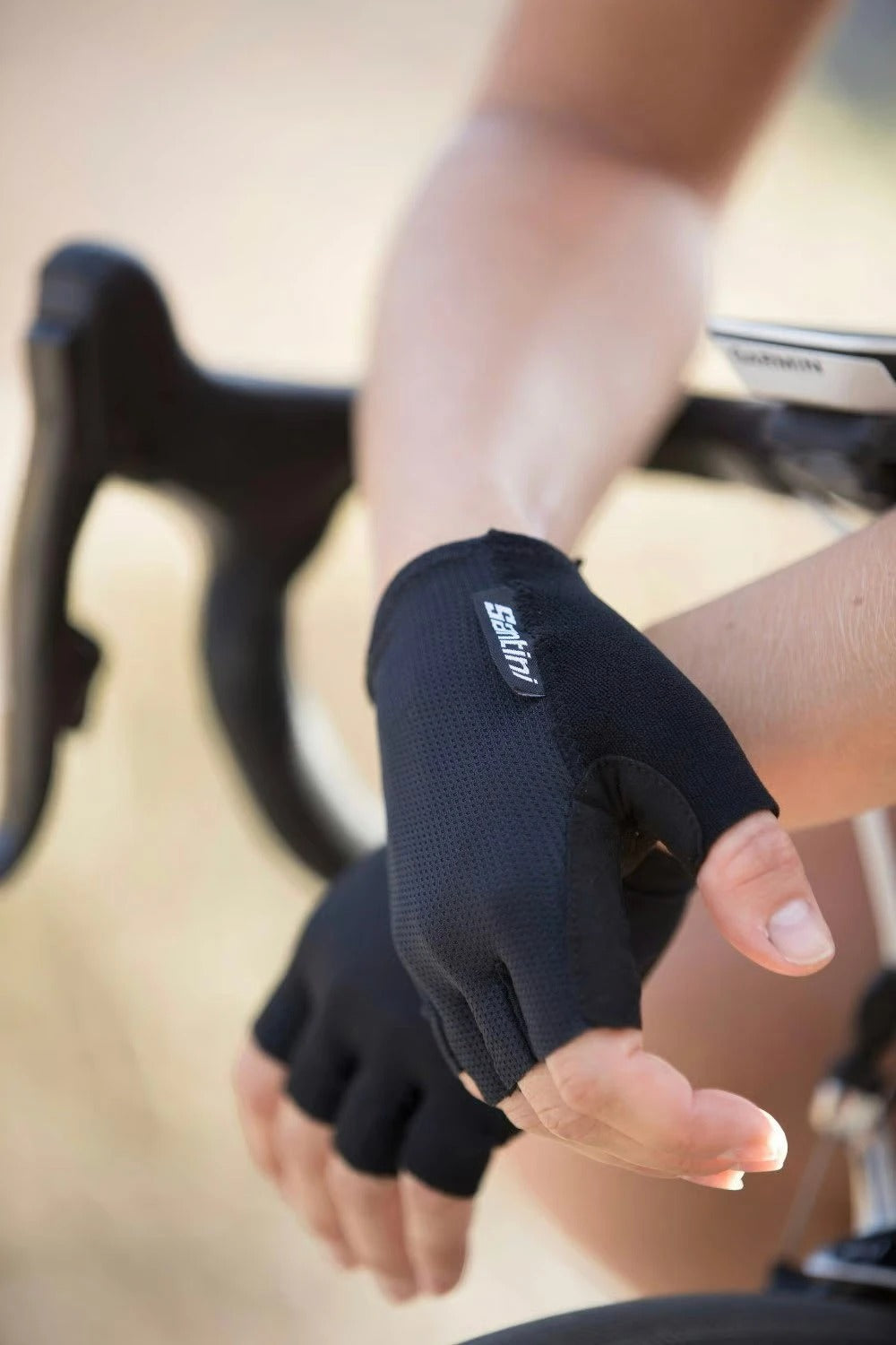 A cyclist leaning on a road bike handlebar wearing black Santini Cycling Gloves with a blurred background