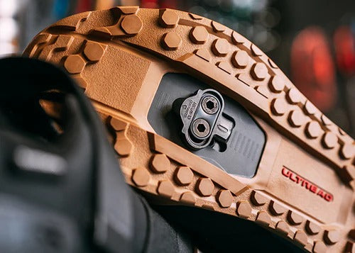 Close-up of the underside of a mountain bike she with a tan sole showing a Shimano SPD cleat against a blurred background