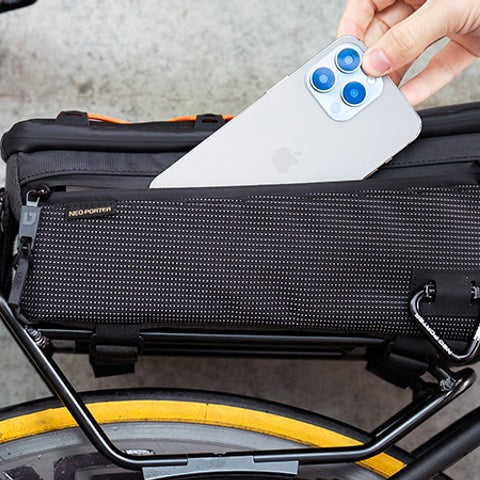 A person storing phone into the side pocket of a black pannier bag attached to bike with a concrete wall in the background