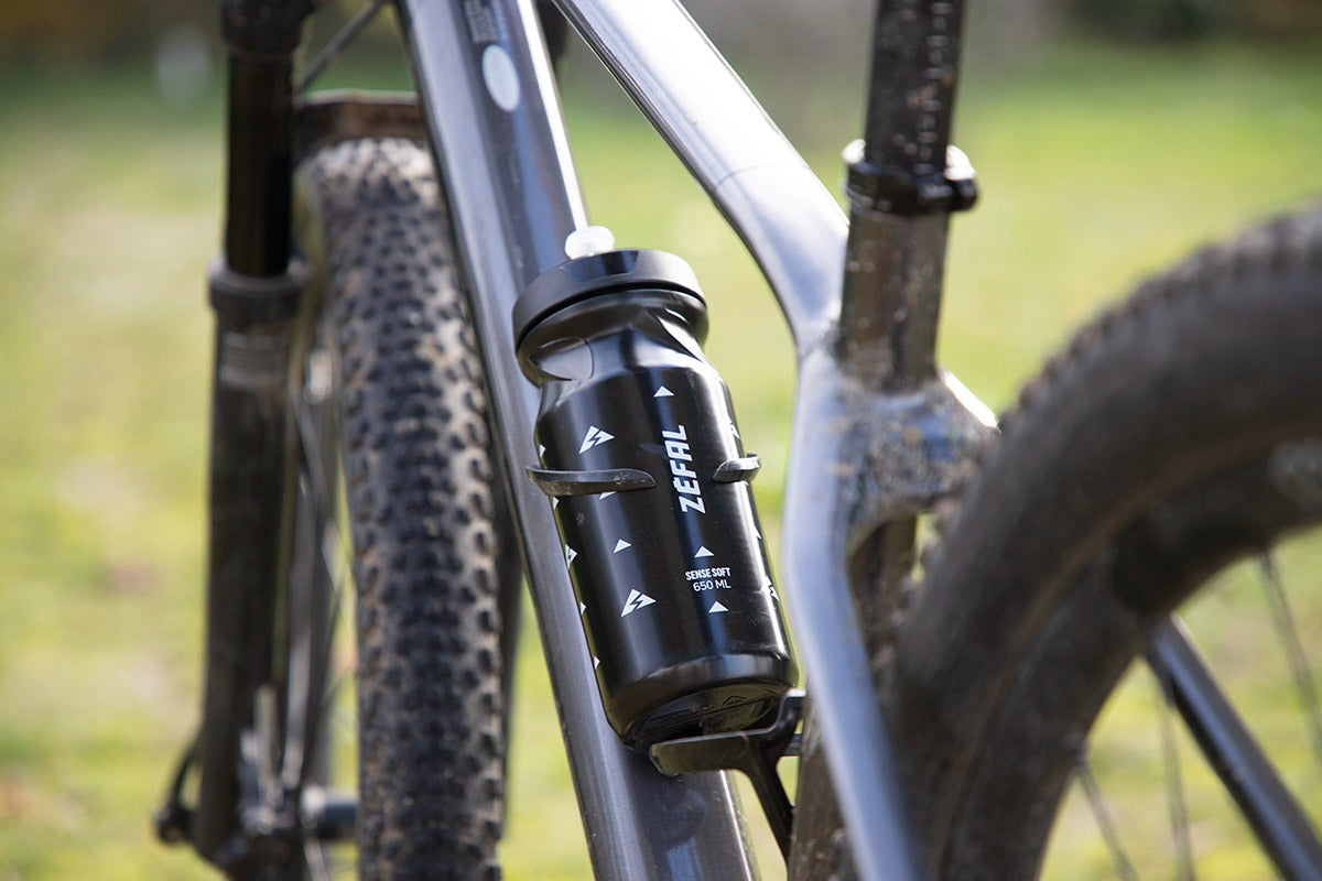 Close-up of a bottle sitting in a bottle cage on a mountain bike with 'Zefal' branding against a blurred natural background.