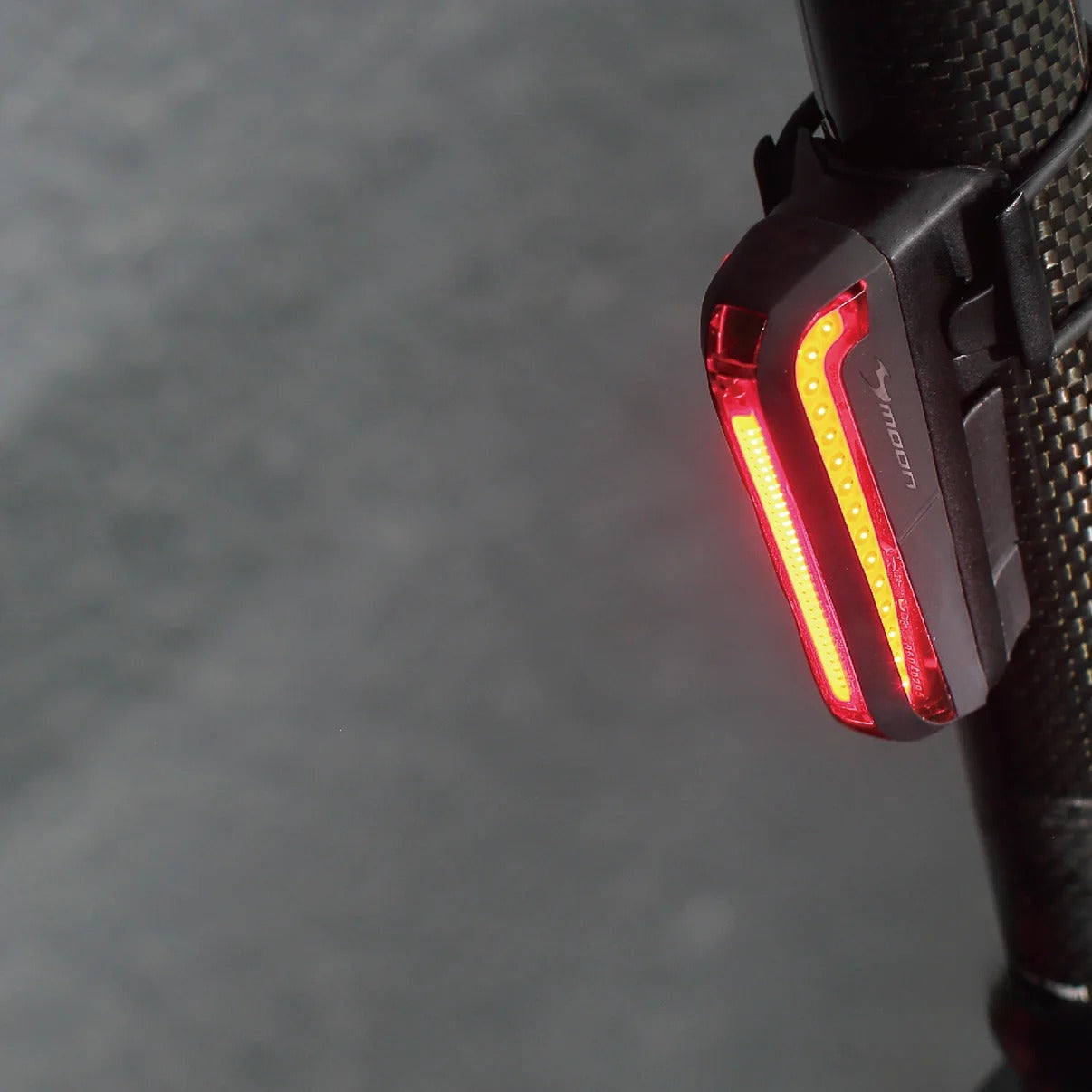 Close-up of a Moon black bicycle rear light with illuminated red LED bars, mounted on a textured black handlebar against a blurred gray background.