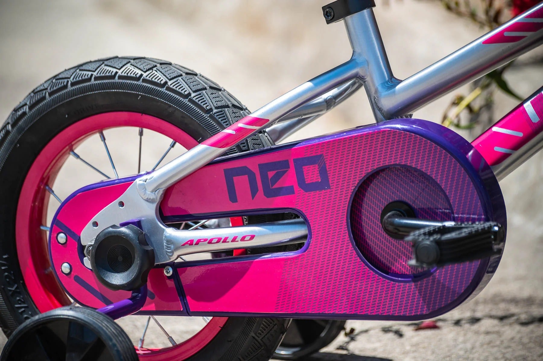 Close-up of a children's Neo bicycle chain guard and frame, featuring a metallic silver frame, a bright pink chain guard with purple accents, and a pink wheel rim.