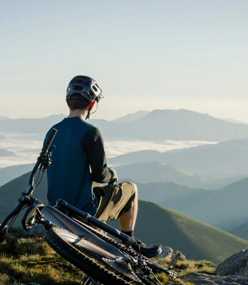 A mountain biker sits on a grassy mountain ridge with a mountain bike in the foreground, looking out over layered blue mountain ranges and a sea of clouds under a pale sky.