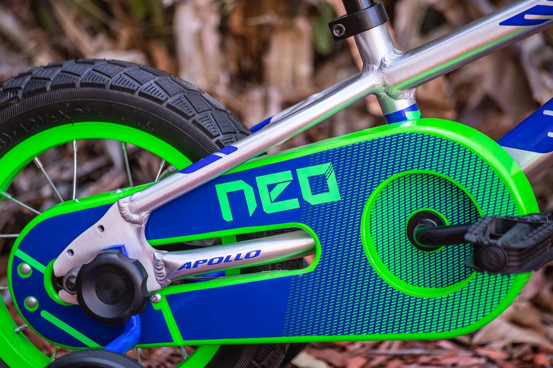 Close-up of a children's bicycle frame and chain guard in bright blue and neon green, with the brand name NEO visible on the chain guard and APOLLO on the chainstay. The bike is photographed outdoors with blurred brown leaves in the background.