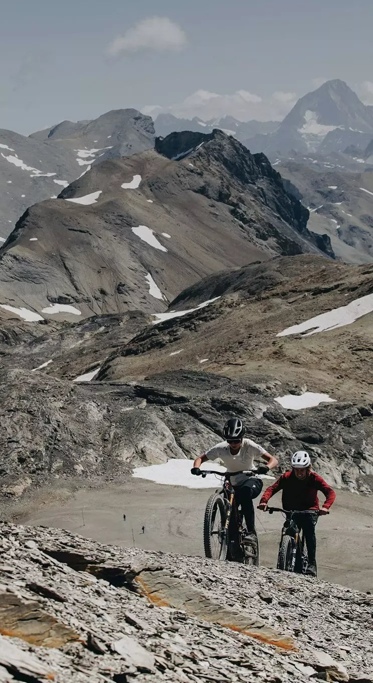 Two mountain bikers on a rocky trail with snow-capped mountains in the background