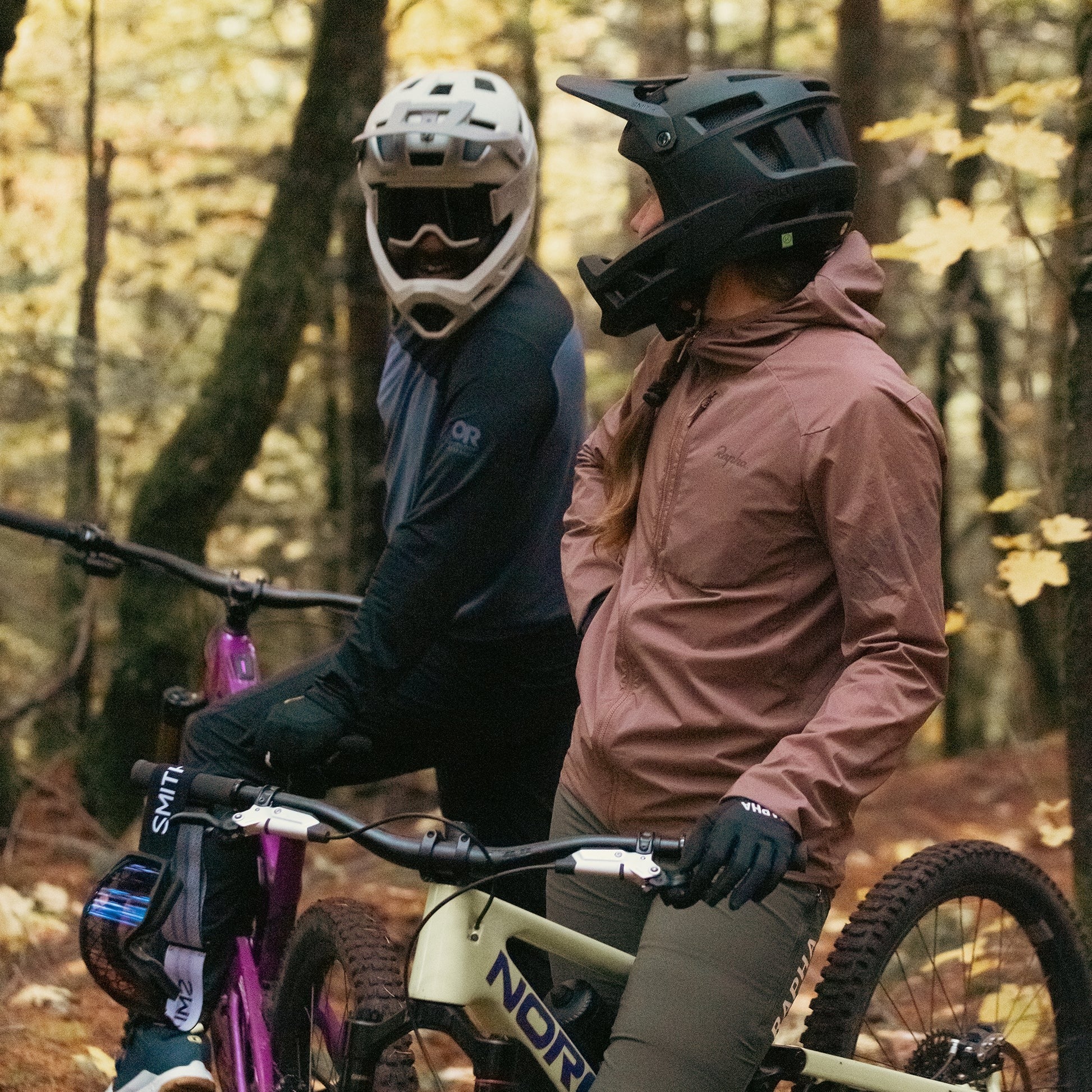 Two mountain bikers in the woods wearing full face mountain bike helmets and riding bikes.