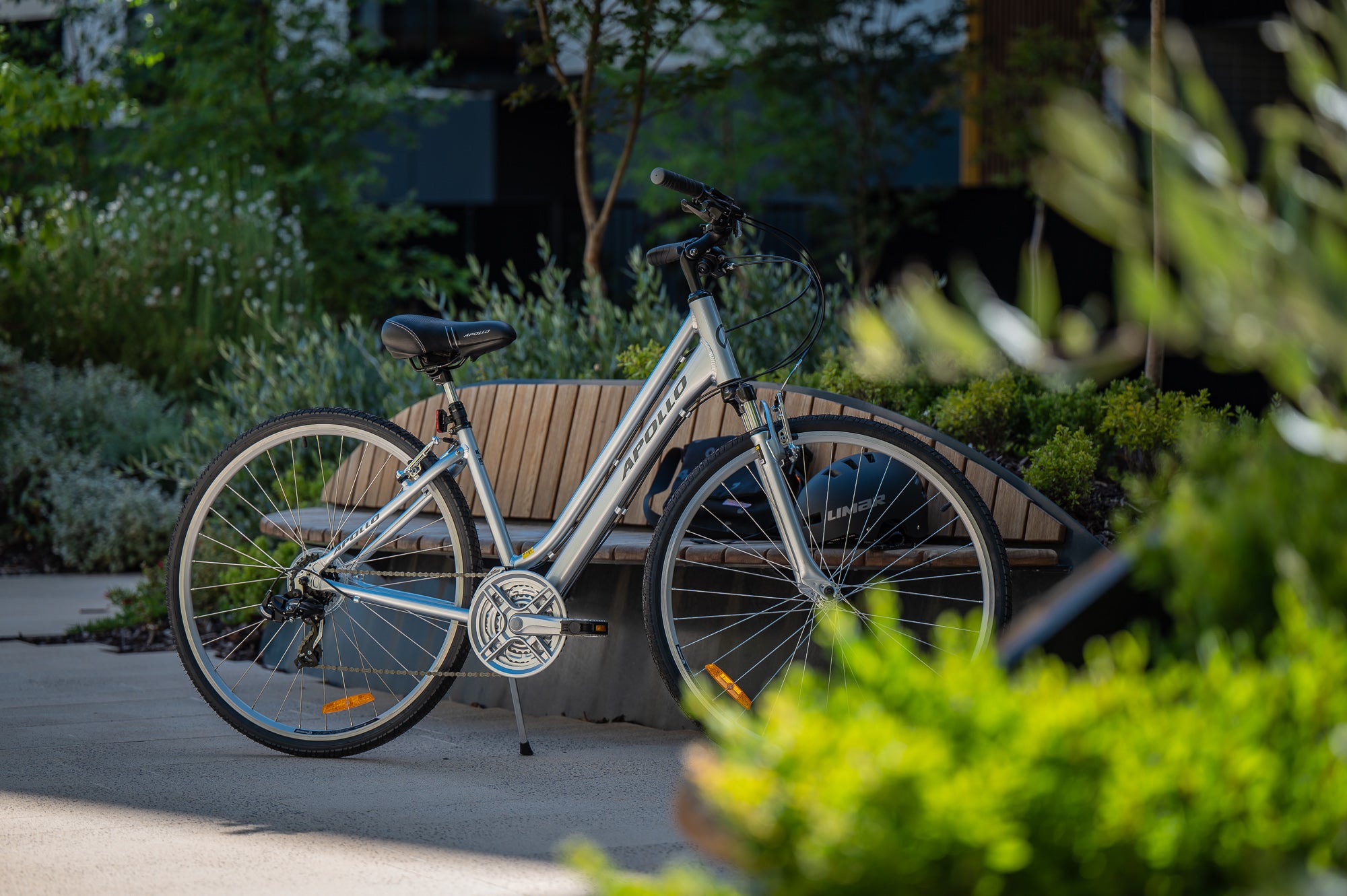Silver Step Through Apollo Hybrid bicycle leaning against a wooden bench in a park setting