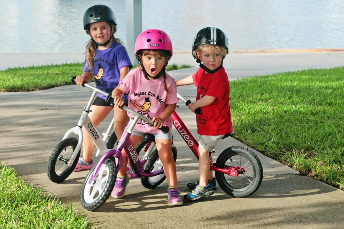 Three children on balance bikes with helmets on a sidewalk.