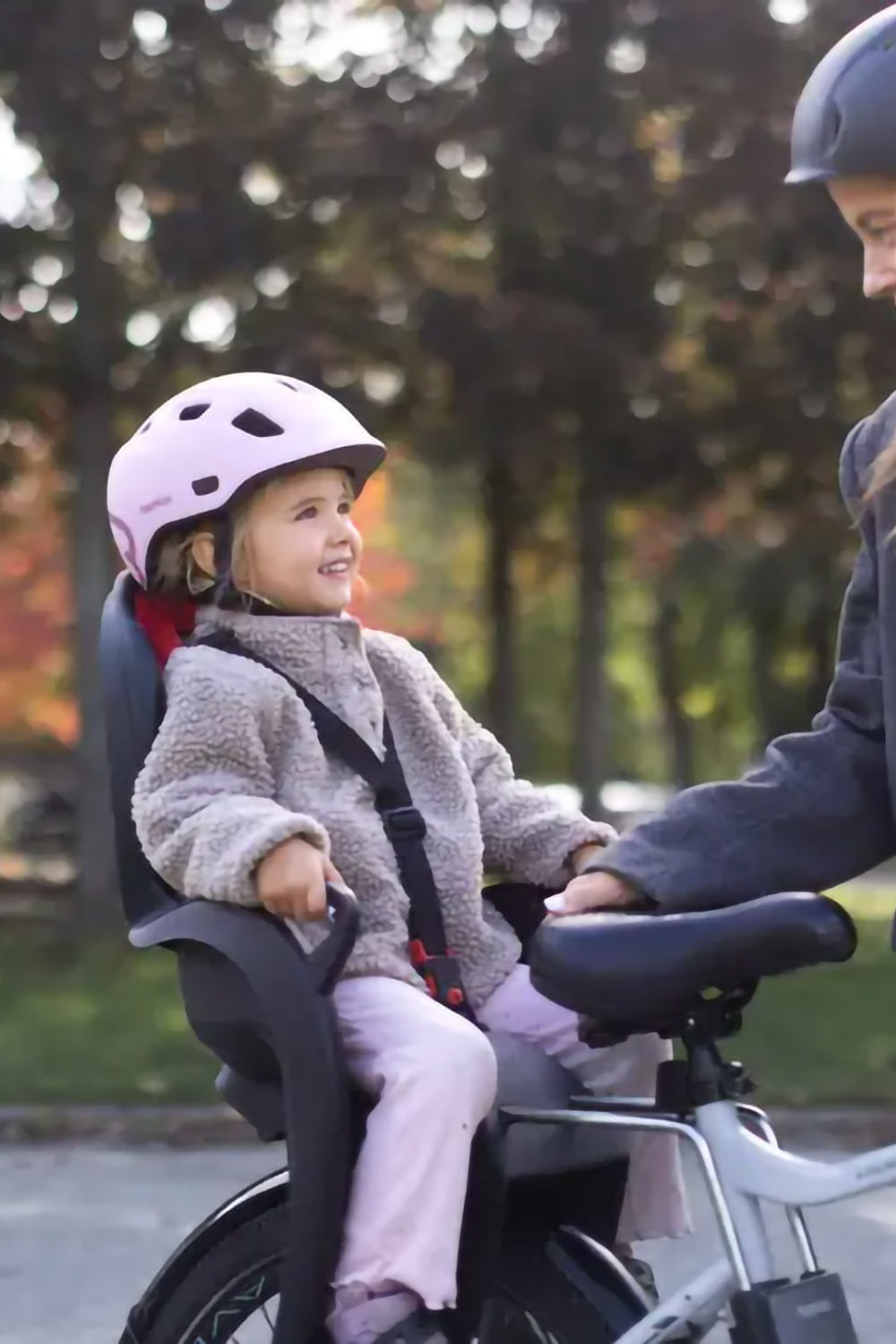 A child in a bicycle baby seat mounted on a bike wearing a pink helmet with a blurred nature background