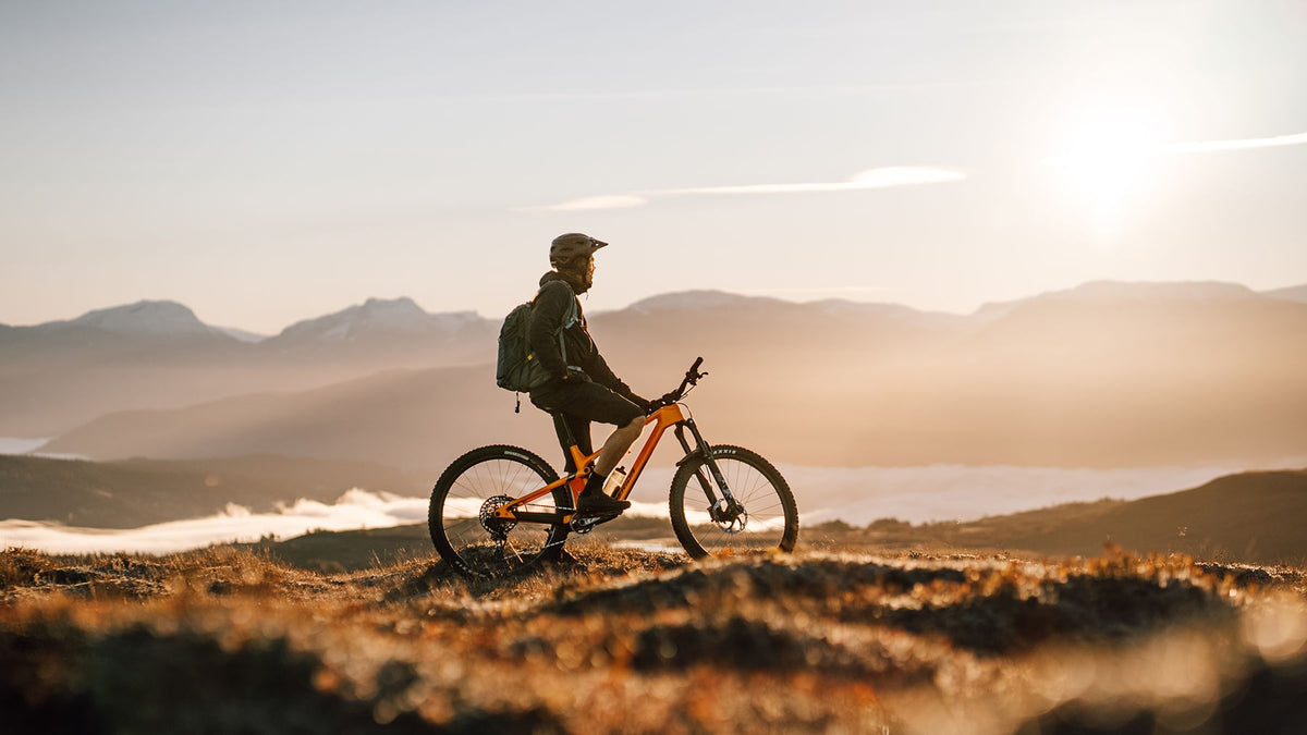 A mountain biker sitting on an orange dual suspension mountain bike against a mountainous background watching the sunset