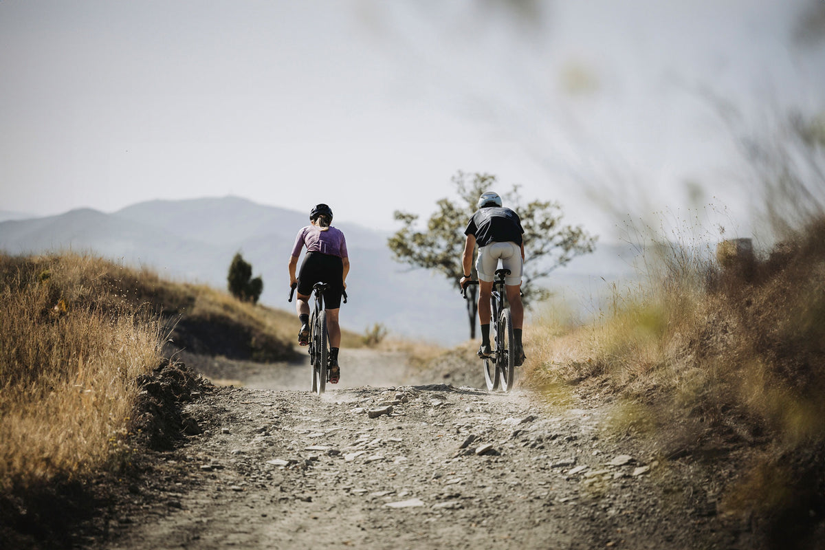 A rear view of two people riding gravel bikes on a dirt road in the country
