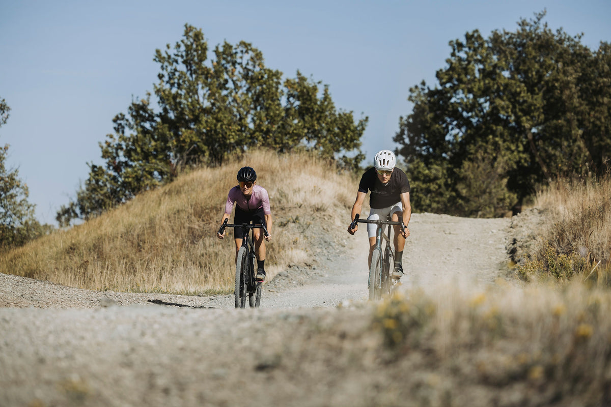 A man and a woman riding gravel bikes on a dirt country road with trees in the background