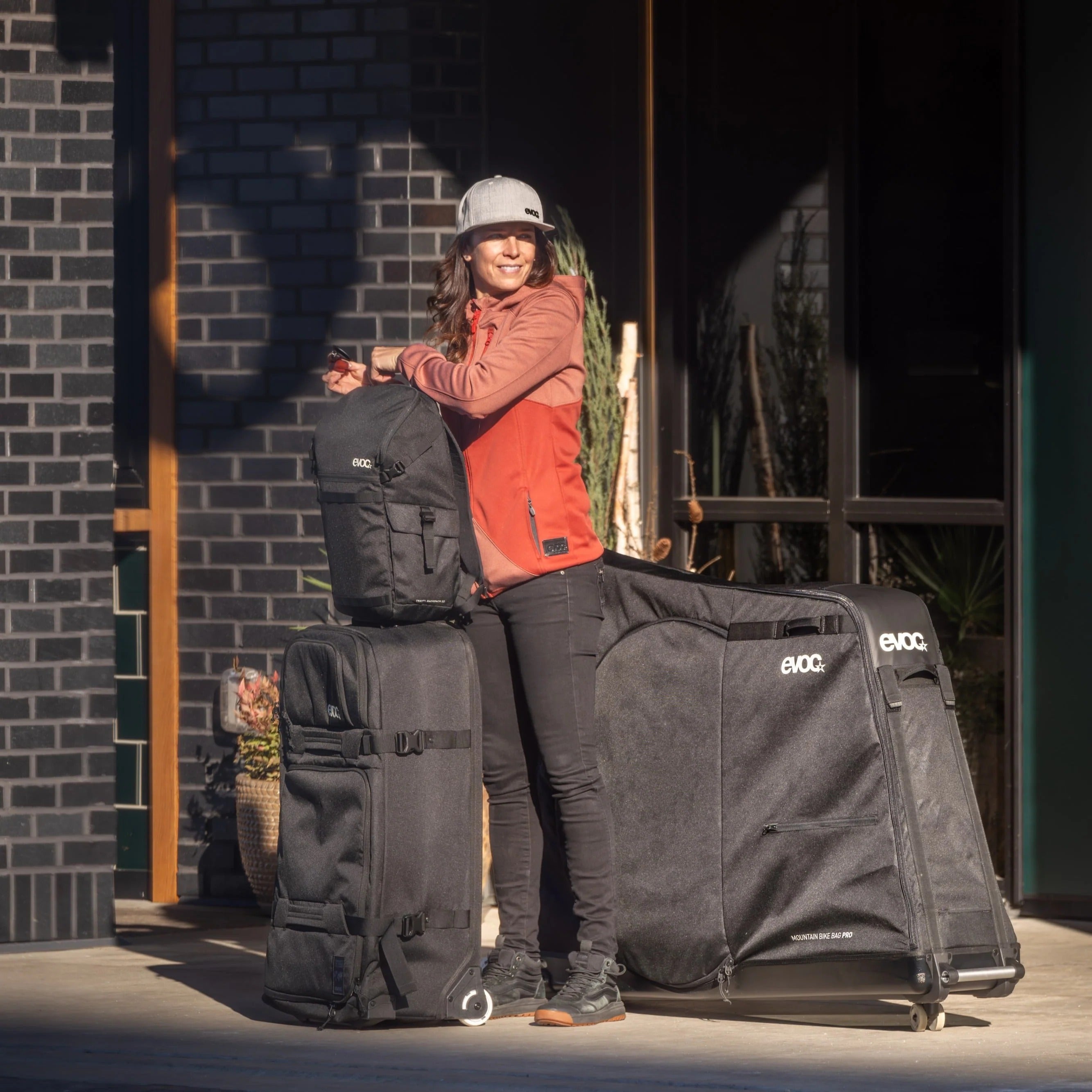Person standing with outdoor gear including a backpack and EVOC bags against a brick wall.