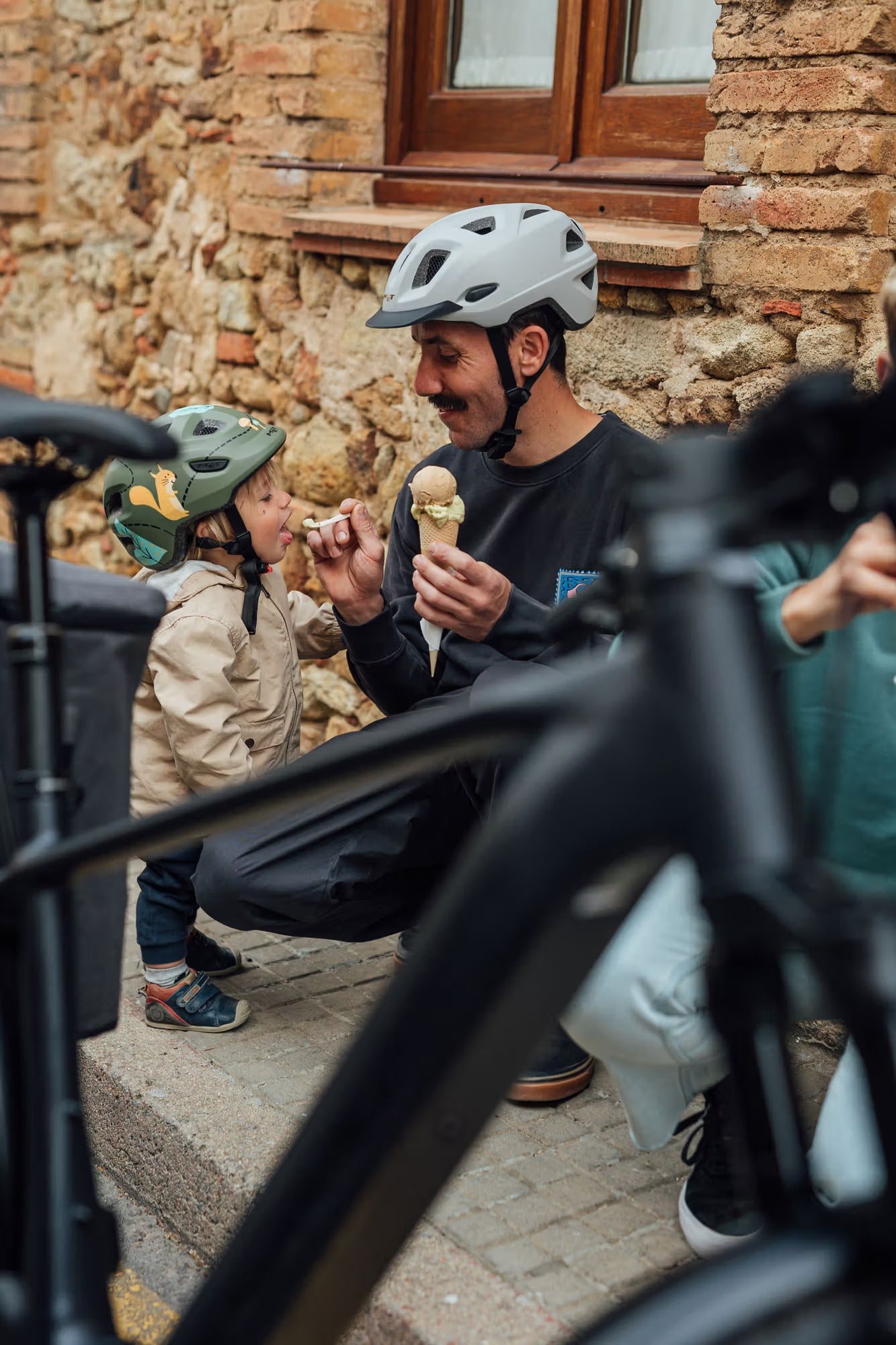 Man and child with helmets sitting on steps next to a bicycle, eating ice cream.