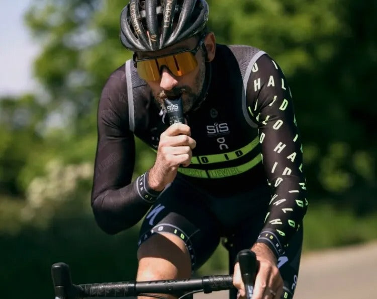Cyclist in a black and green outfit with a helmet and sunglasses, sucking on a SIS energy gel while riding on a road bike outdoors.