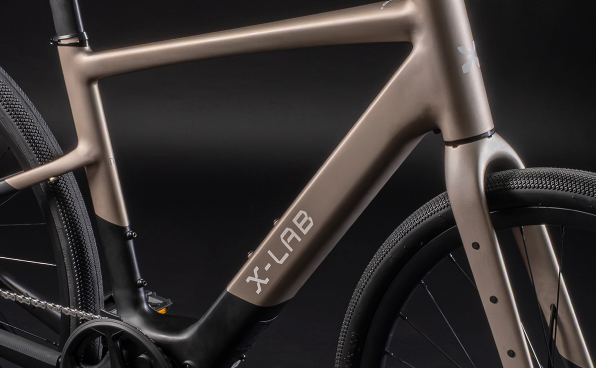 Close-up of a bicycle frame in a matte taupe metallic finish with black components, featuring the X-LAB brand name on the down tube against a dark studio background.