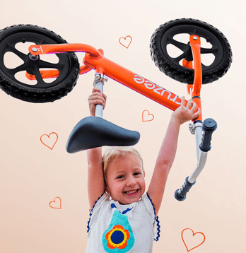 Child holding an orange balance bike against a beige background with heart shapes