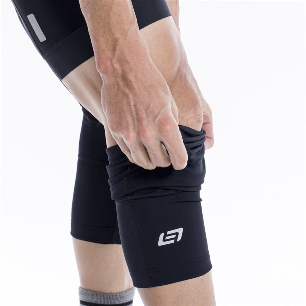 Person wearing black cycling shorts and putting on black cycling knee sleeves with a brand logo on a white background