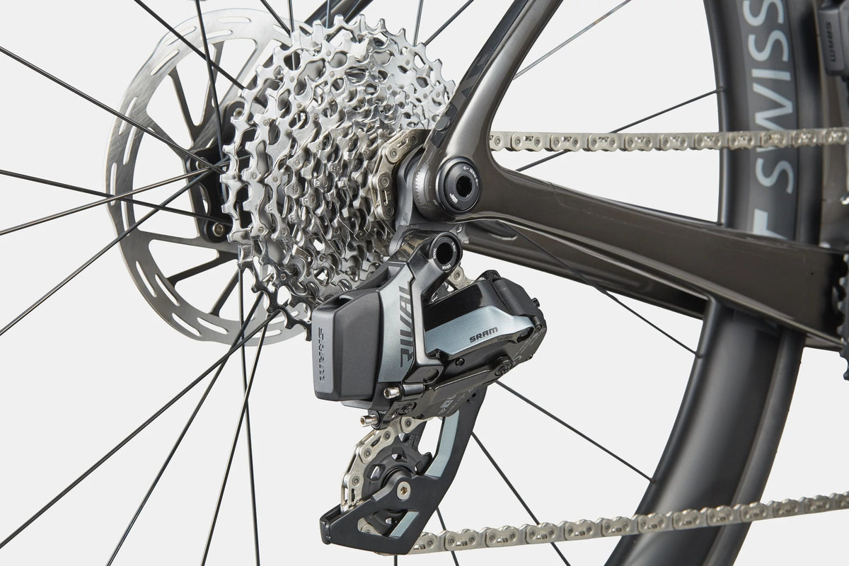 Close-up of a bicycle rear derailleur and cassette, showing an electronic SRAM Rival rear derailleur mounted on a charcoal road bike frame with a disc brake and chainring.