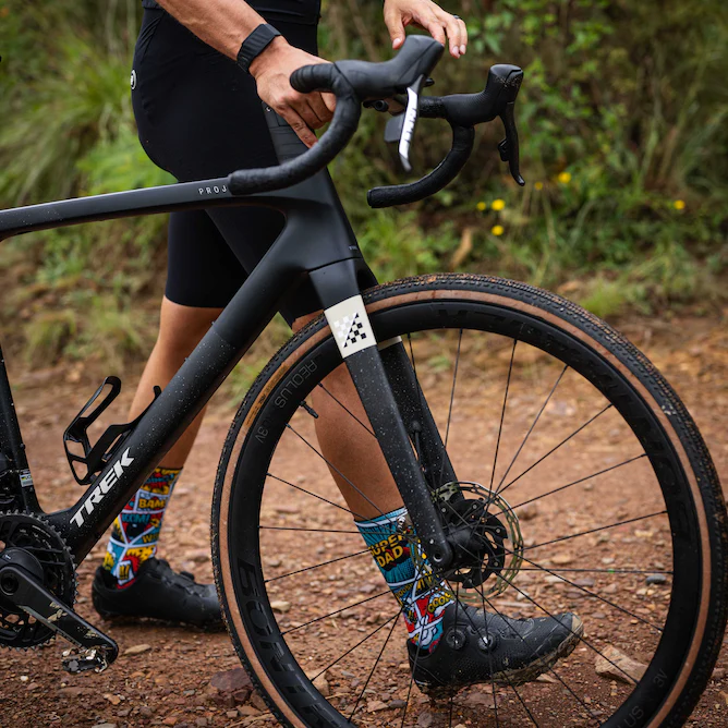 A close-up outdoor shot of a cyclist's legs and hands on a black Trek road bike, with the rider wearing colorful patterned socks and black cycling shoes on a dirt trail.