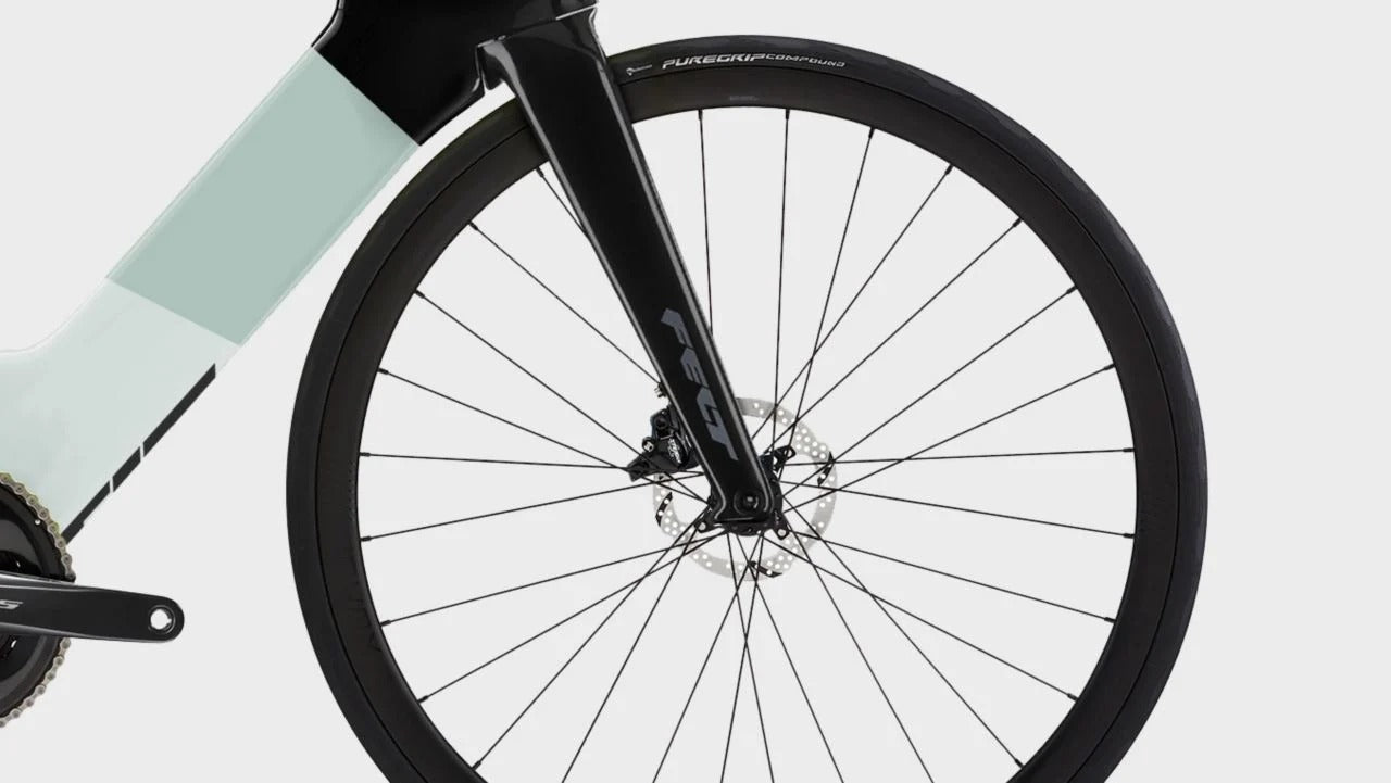 Close-up of a bicycle wheel and front fork with a pale mint green frame section, black rim, and visible Shimano components on a light gray background.