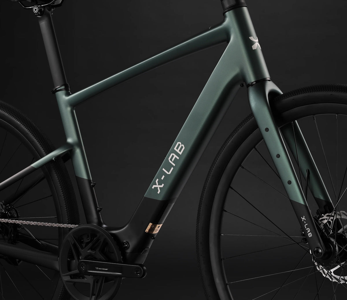 Close-up studio shot of a bicycle frame and front wheel against a dark background. The frame is matte dark green with black components and visible X-LAB branding on the down tube and fork.