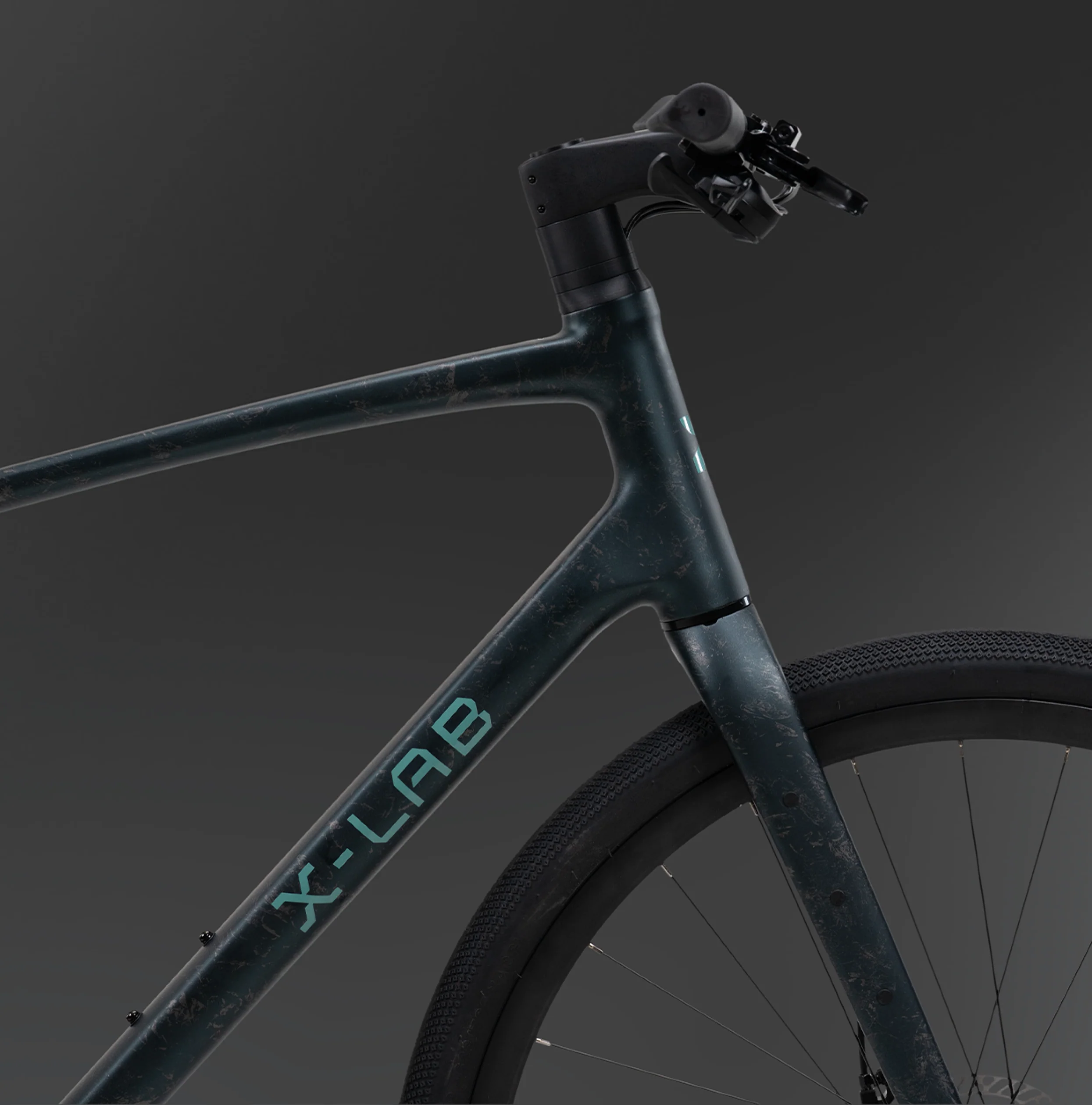 Close-up studio shot of an X-Lab Hybrid Bike and handlebar area with part of the front wheel visible, on a dark gray gradient background. The frame has a dark metallic teal finish with a subtle distressed texture and the brand name X-LAB printed in light teal on the down tube.