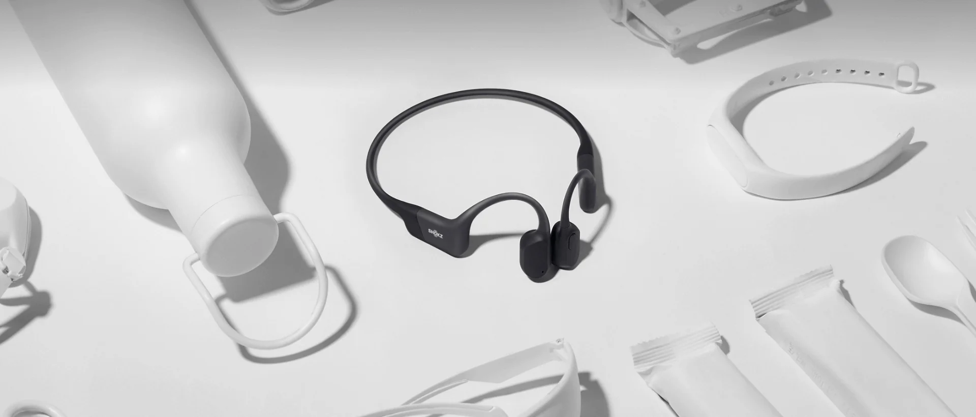 Black wireless earbuds on a white surface with blurred electronic devices in the background