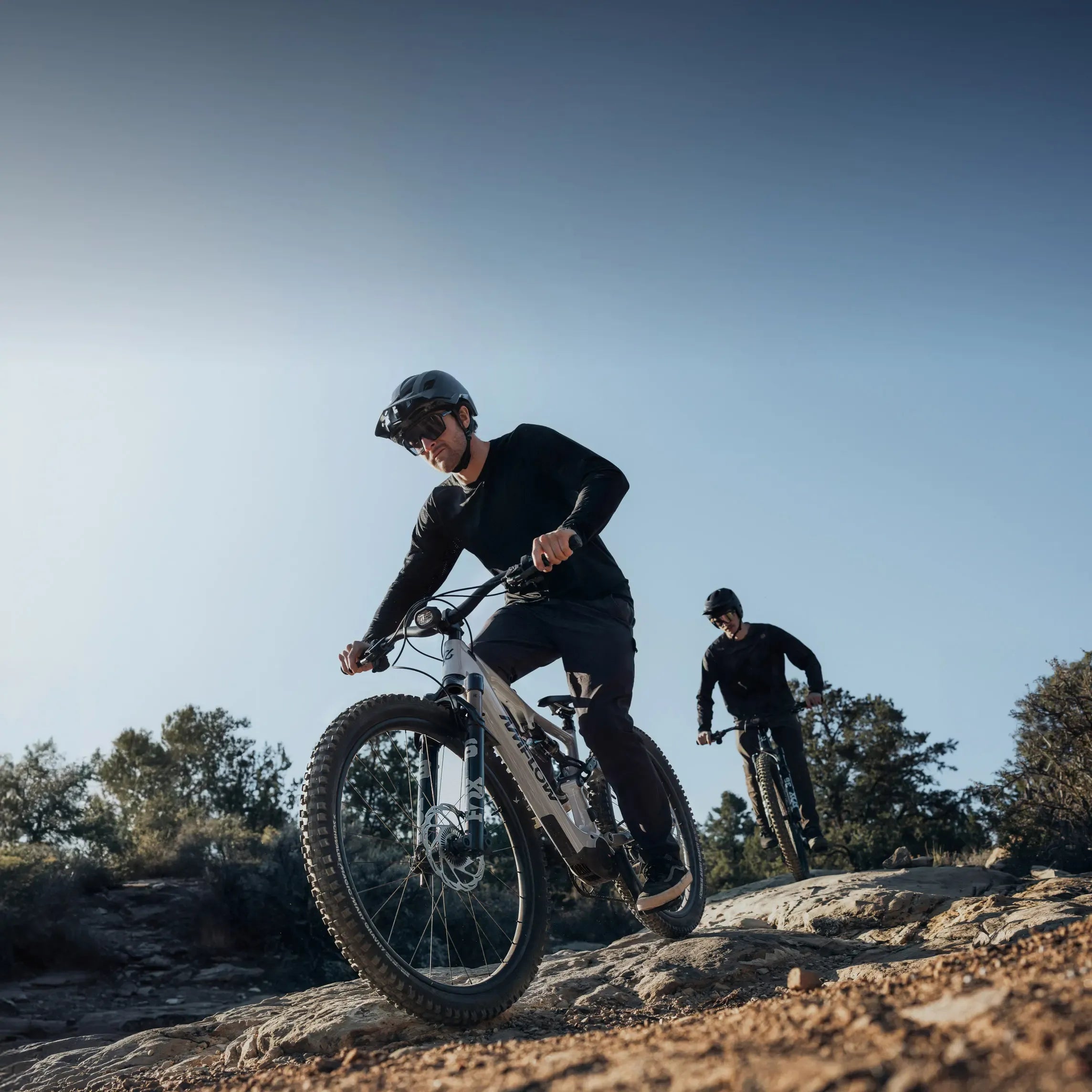 Two mountain bikers riding on a rocky trail with a clear blue sky.