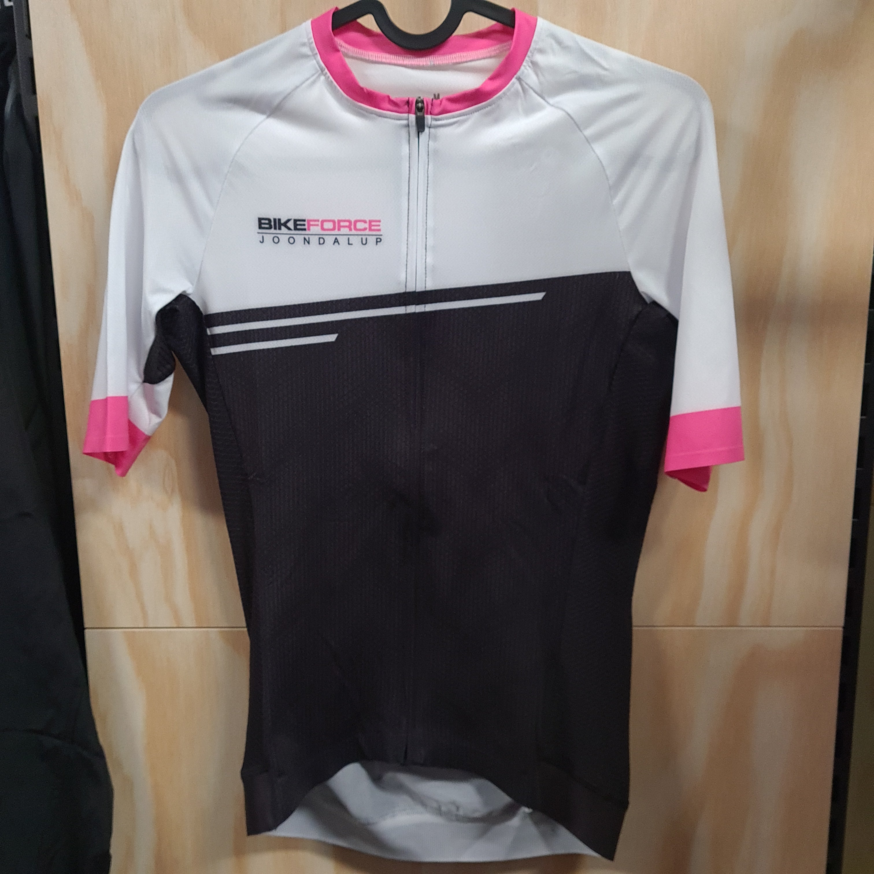 A short-sleeve cycling jersey hanging on a black hanger against a light wood panel background. The jersey has a white upper section, a black lower section, and bright pink accents on the collar, sleeve cuffs, and side panels. The logo 'Bike Force Joondalup' is printed in black and pin on the right upper chest.
