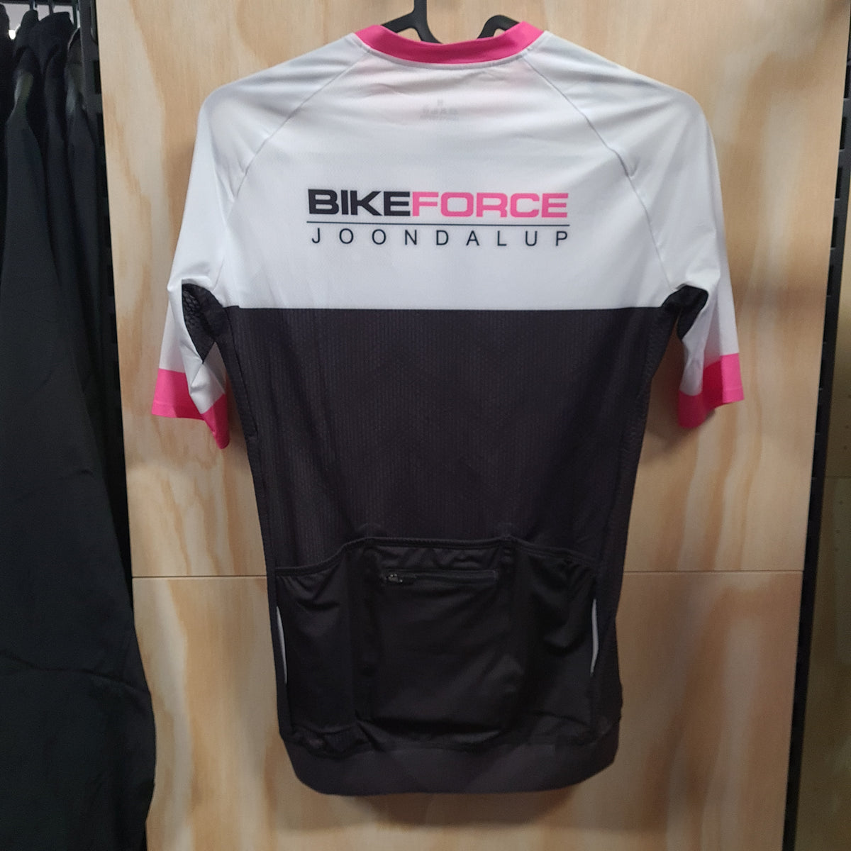 A short-sleeve cycling jersey hanging on a black hanger against a light wood-paneled wall. The jersey is white on the upper chest and shoulders, black on the lower body and sleeves, with pink accents on the collar, sleeve cuffs, and the 'BIKEFORCE JOONDALUP' text across the upper back.