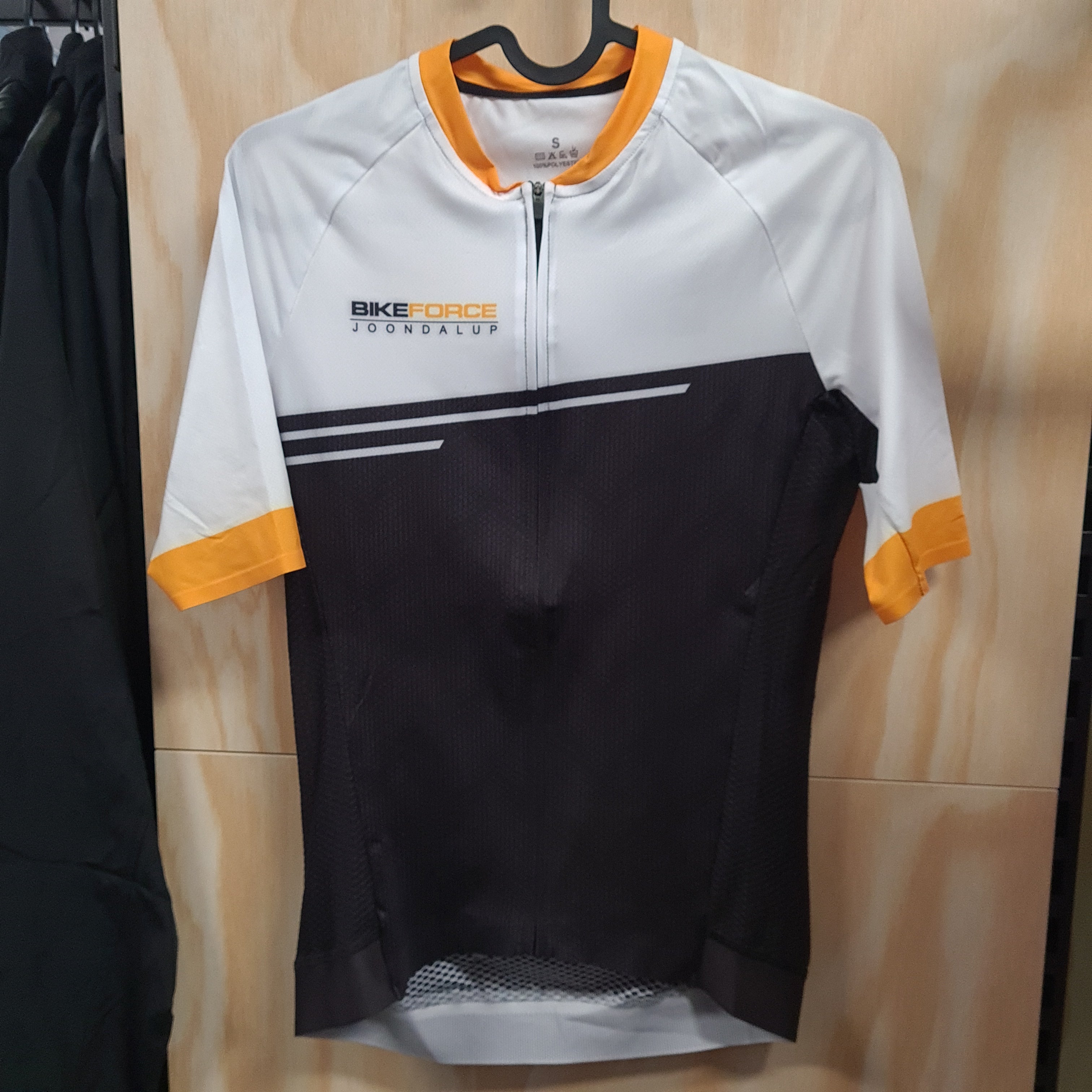 A short-sleeve cycling jersey hanging on a black hanger against a light wood-paneled wall. The jersey is white on the upper chest and sleeves, black on the lower torso, and features yellow accents on the collar, cuffs, and Bike Force Joondalup logo printed in yellow and black on the left hand upper chest.