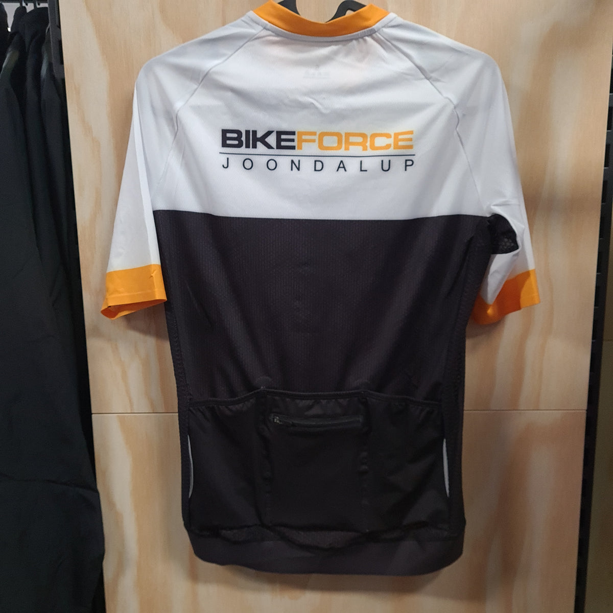 A short-sleeve cycling jersey with a white upper section, black lower section, and yellow sleeve cuffs, featuring the text 'BIKEFORCE JOONDALUP' on the back. The jersey is hanging on a wooden wall.