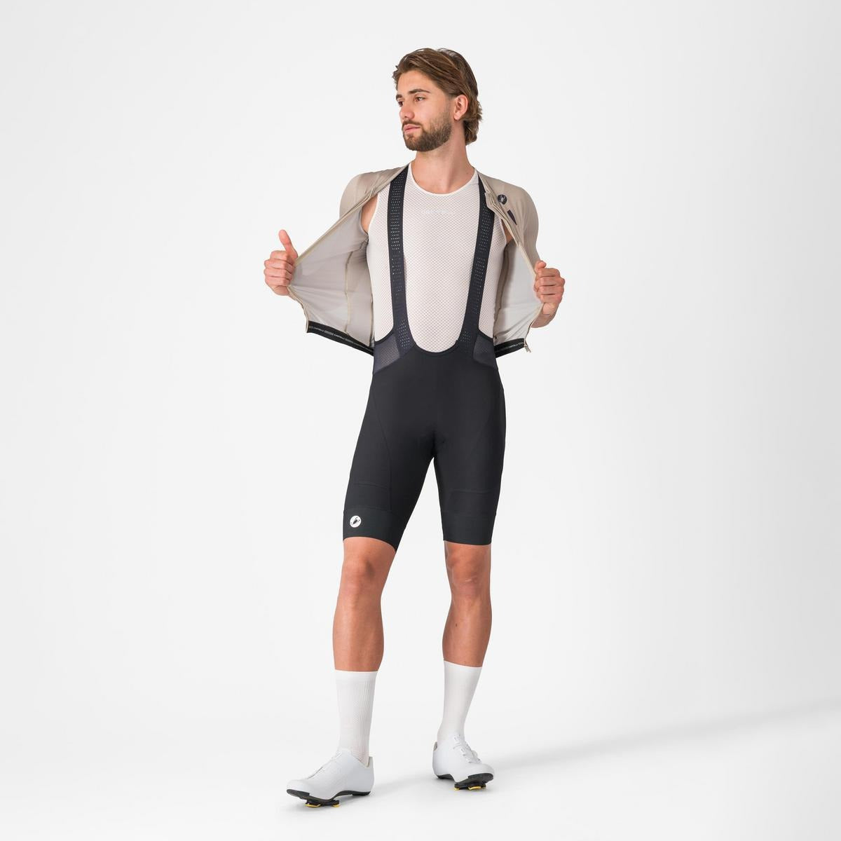 A man wearing a cycling jersey unzipped showing the black bibshorts underneath on a light grey background