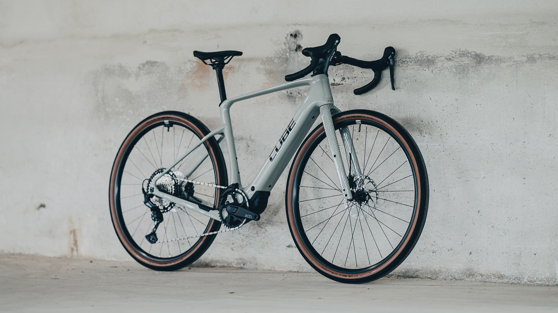 Beige Cube Gravel Bike with black components leaning against a light gray wall.