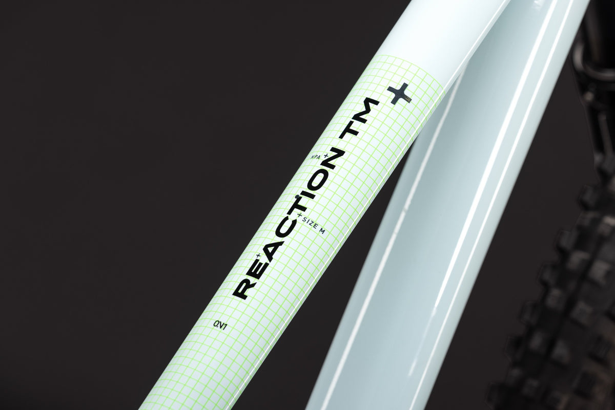 Close-up of a bicycle frame tube with a pale mint green grid-patterned decal reading REACTION MT+ and small model text, set against a dark background.