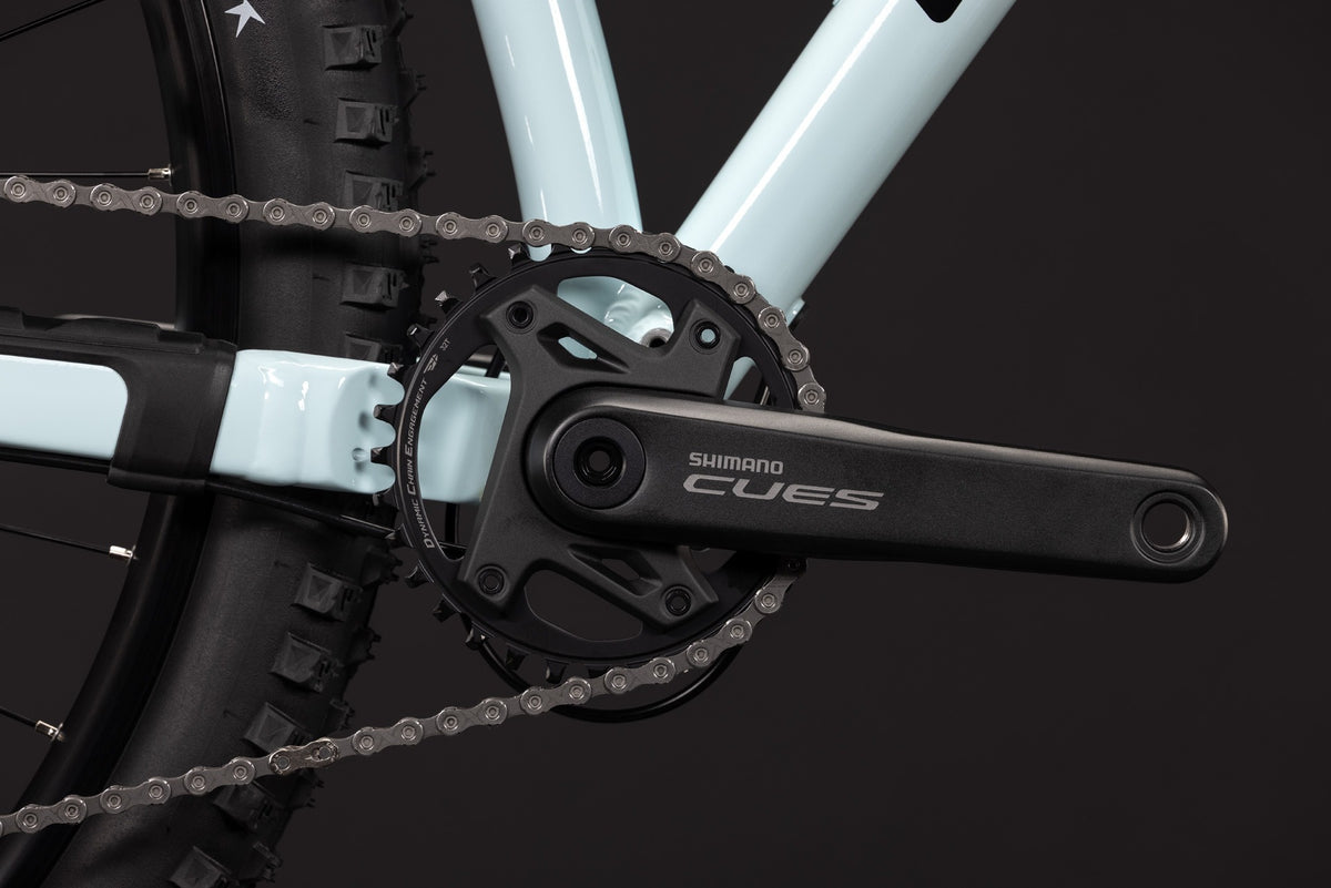 Close-up of a Shimano Cues bicycle crankset with a black chainring and black crank arm, attached to a light blue bicycle frame against a dark background.